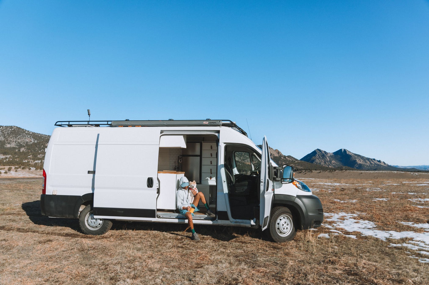 A person and their dog relax in a white camper van, an RV rental, with doors open, parked in a scenic mountain field.