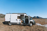 A person and their dog relax in a white camper van, an RV rental, with doors open, parked in a scenic mountain field.