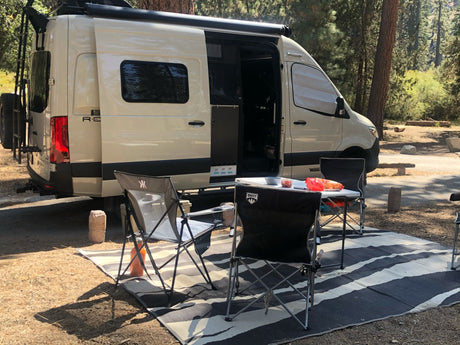 RV rental camper van at a wooded campsite, with an outdoor area set up for relaxing and enjoying the outdoors.