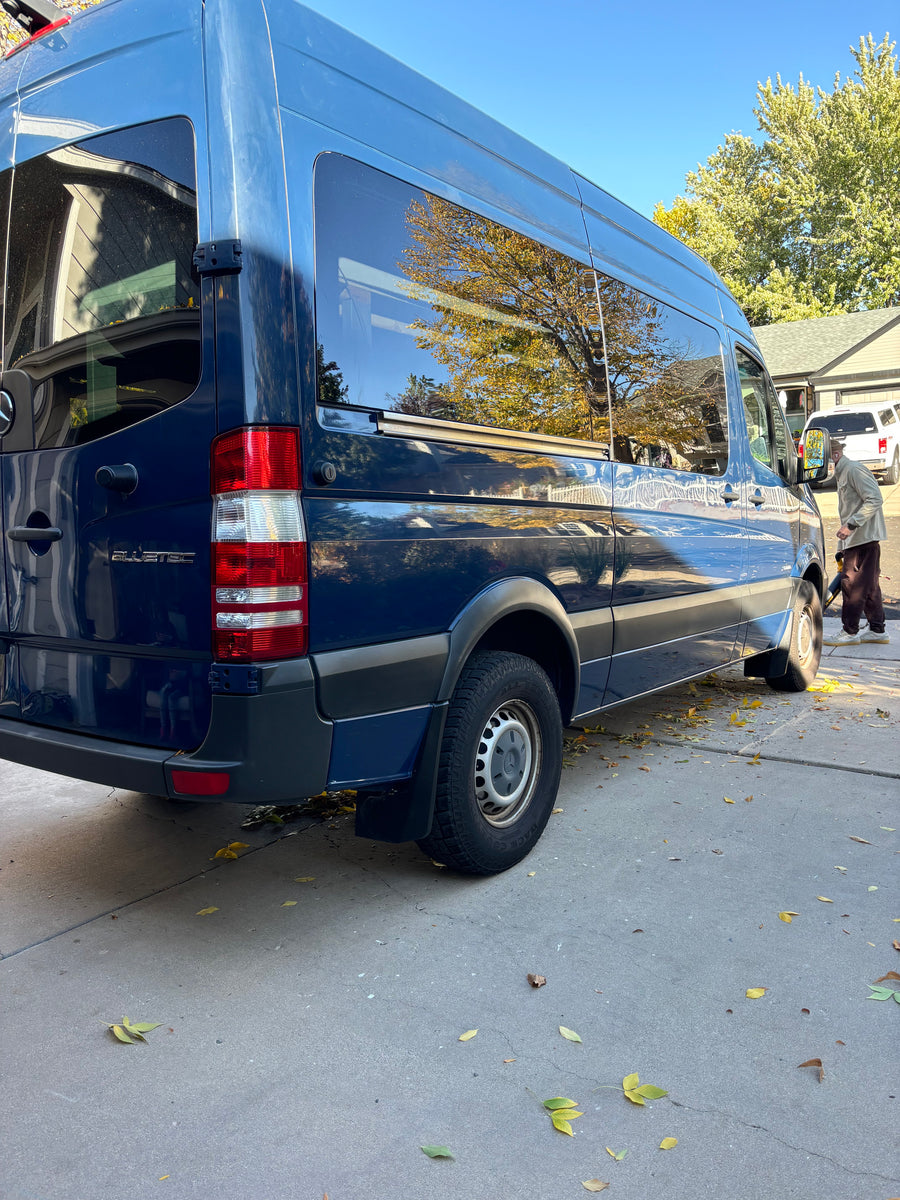 Dark blue Mercedes Sprinter van, perfect for an rv rental, parked on a driveway with autumn leaves. A person clears leaves.