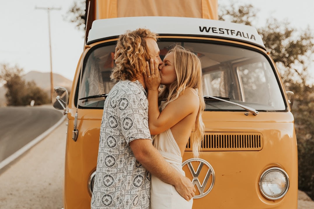 A loving couple kisses by their orange VW Westfalia camper van, perfect for an RV rental road trip adventure!