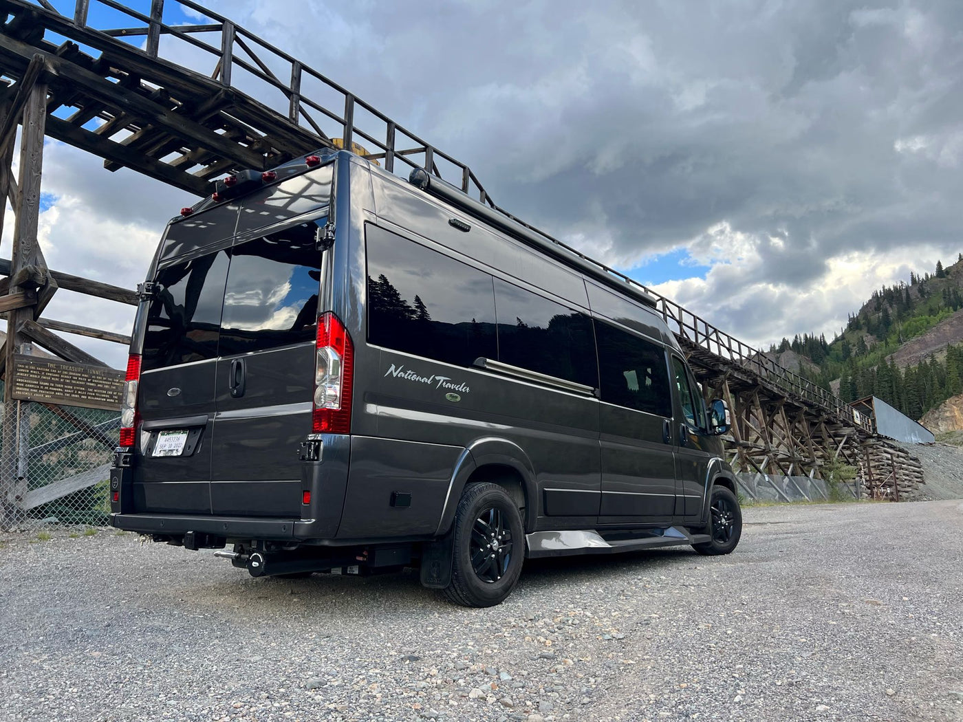  with red-streaked mountains under cloudy skies.|RV rental parked near historic Treasury Tunnel trestle. Explore mountains & adventure in this National Traveler van.|Dark camper van (RV rental) by old wooden trestle — photo 2