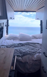 Inside an RV rental, a cozy bed faces open doors revealing a panoramic blue ocean and sky view. Perfect for scenic travel!