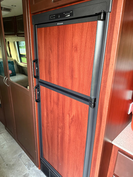 A Dometic RV refrigerator with wood grain doors and control panel inside an RV rental. Reflection shows the vehicle interior.