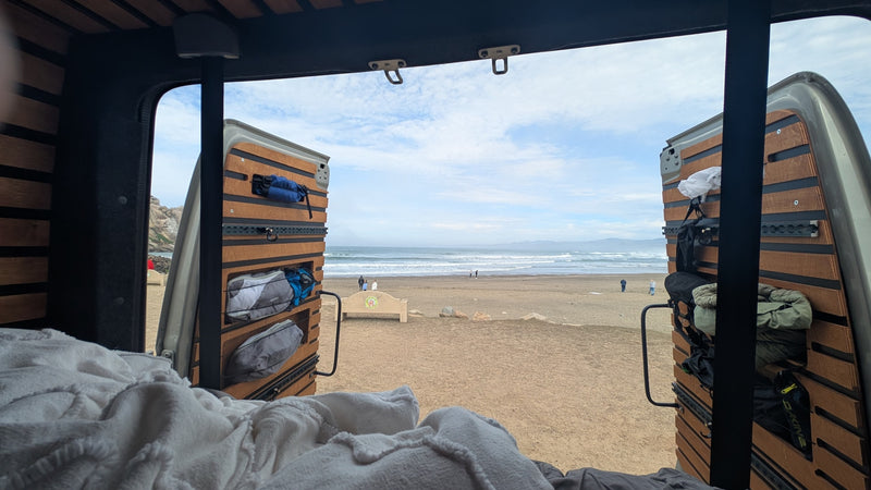 Cozy rv rental view from bed: ocean waves, sandy beach, and distant hills seen through open camper van doors. — photo 11