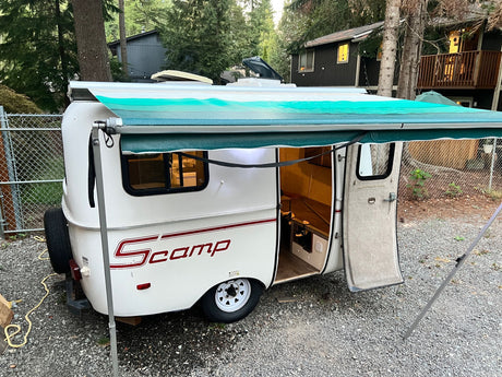 White Scamp compact travel trailer with extended awning, parked on gravel. Great for an RV rental getaway!