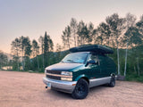 Green Astro van with rooftop tent parked on a dirt road by a lake. An ideal RV rental for outdoor journeys.