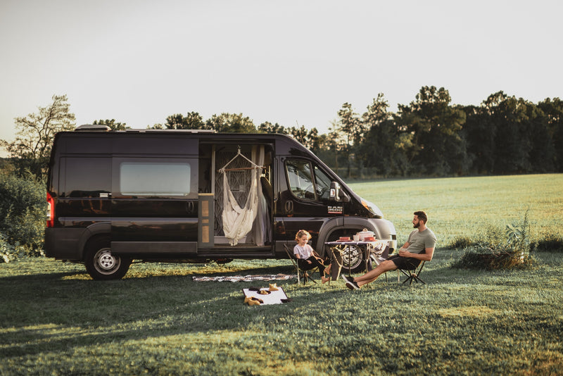 RV rental getaway: A man & child share a meal outside their black camper van in a serene grassy field. Perfect for family camping. — photo 15