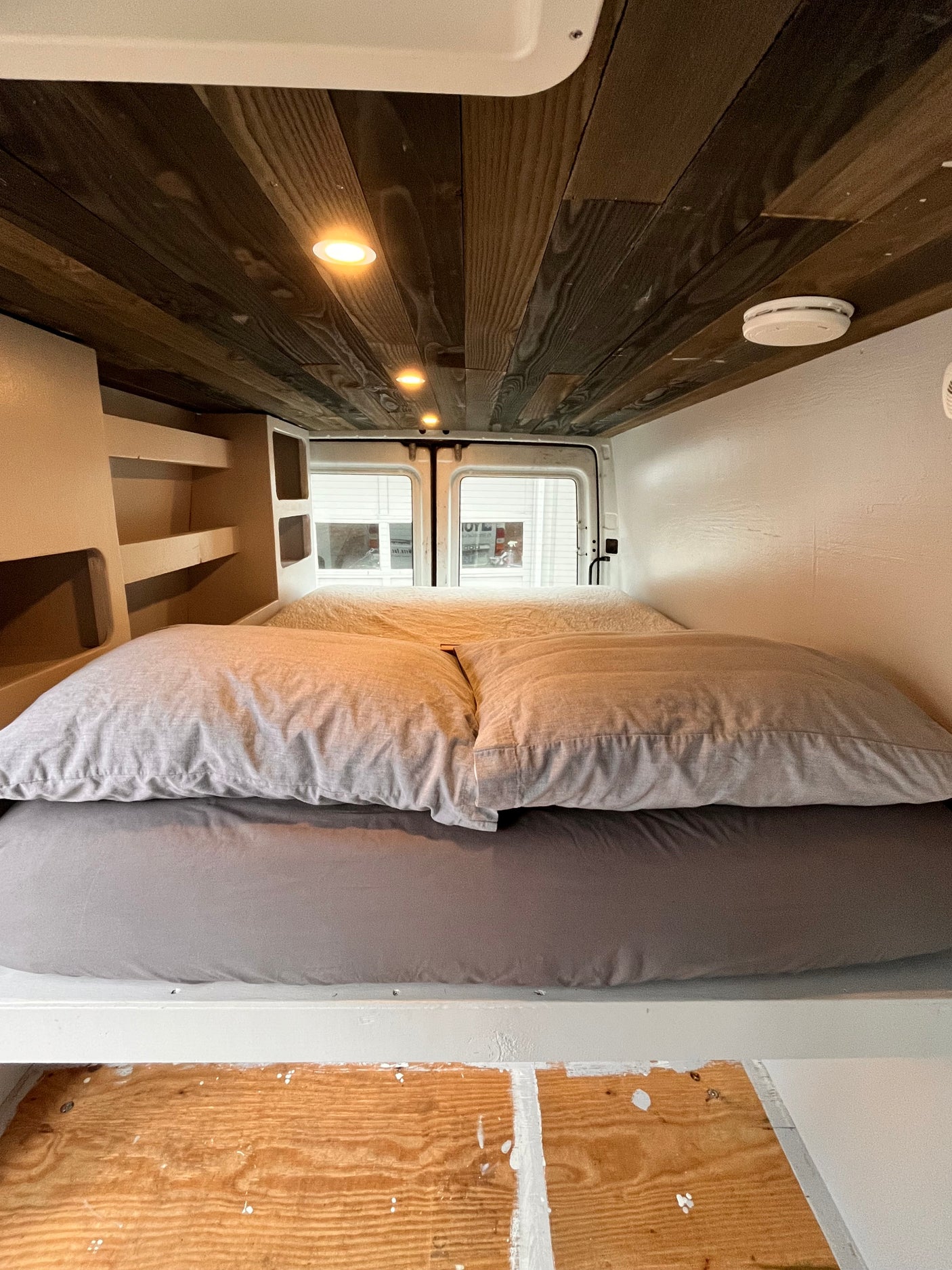 Inside an RV rental: a snug bed with gray bedding, built-in shelves, and a rustic wooden ceiling with lights.