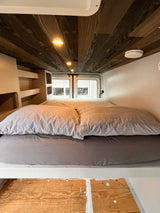Inside an RV rental: a snug bed with gray bedding, built-in shelves, and a rustic wooden ceiling with lights.