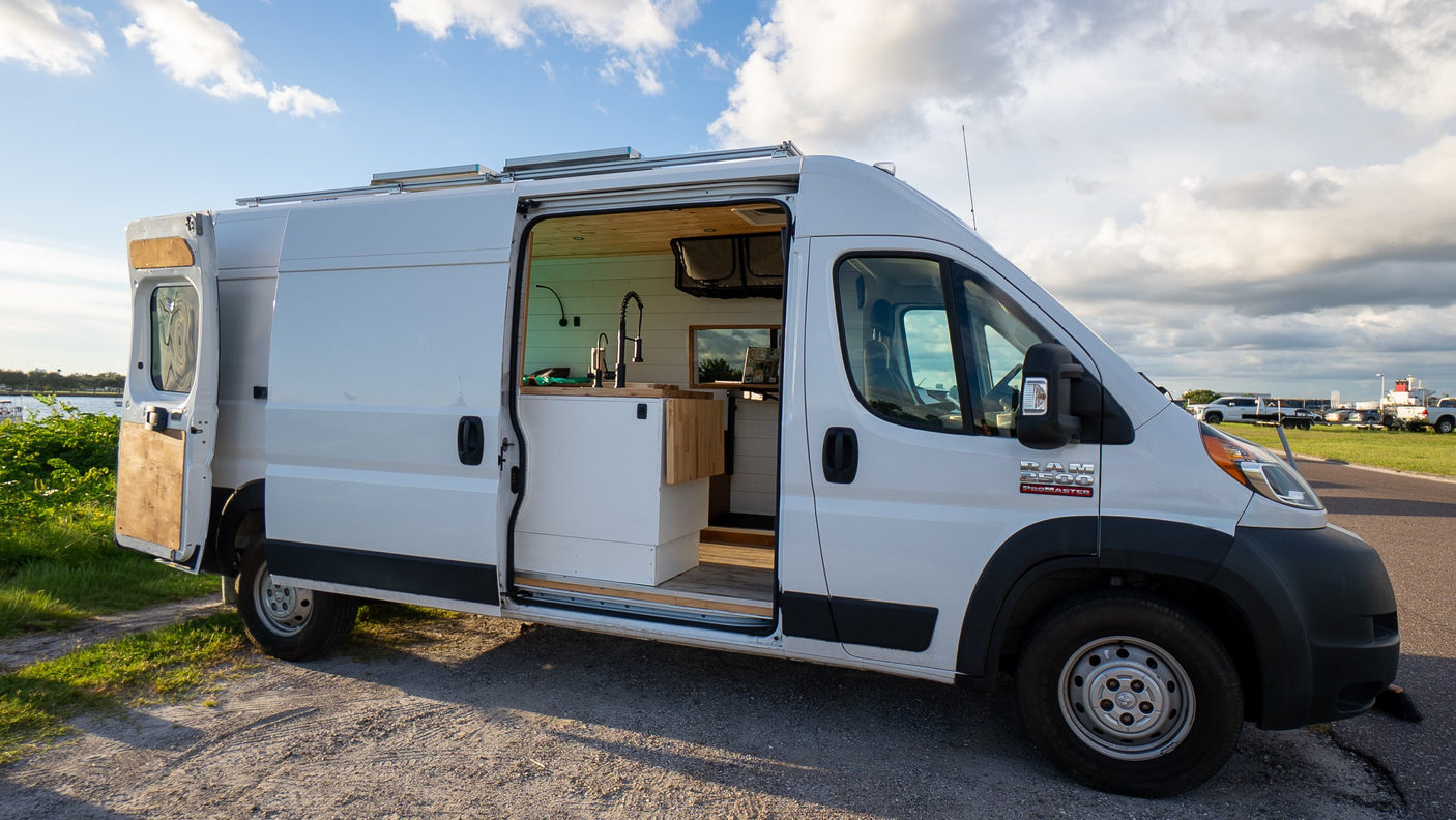 White Ram ProMaster rv rental van conversion, side door open to a minimalist kitchen. Parked outside under a cloudy sky.