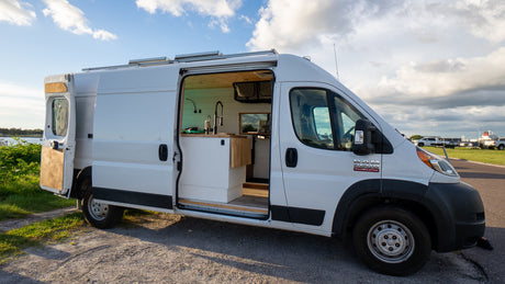 White Ram ProMaster rv rental van conversion, side door open to a minimalist kitchen. Parked outside under a cloudy sky.