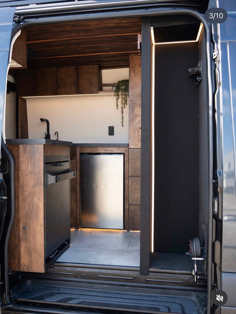 Interior of a modern RV rental van conversion, showing a dark wood kitchenette with a stainless fridge and LED lighting.