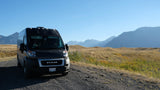 Black RAM camper van, possibly an RV rental, on a gravel road. Panoramic views of mountains & golden hills under a clear sky.