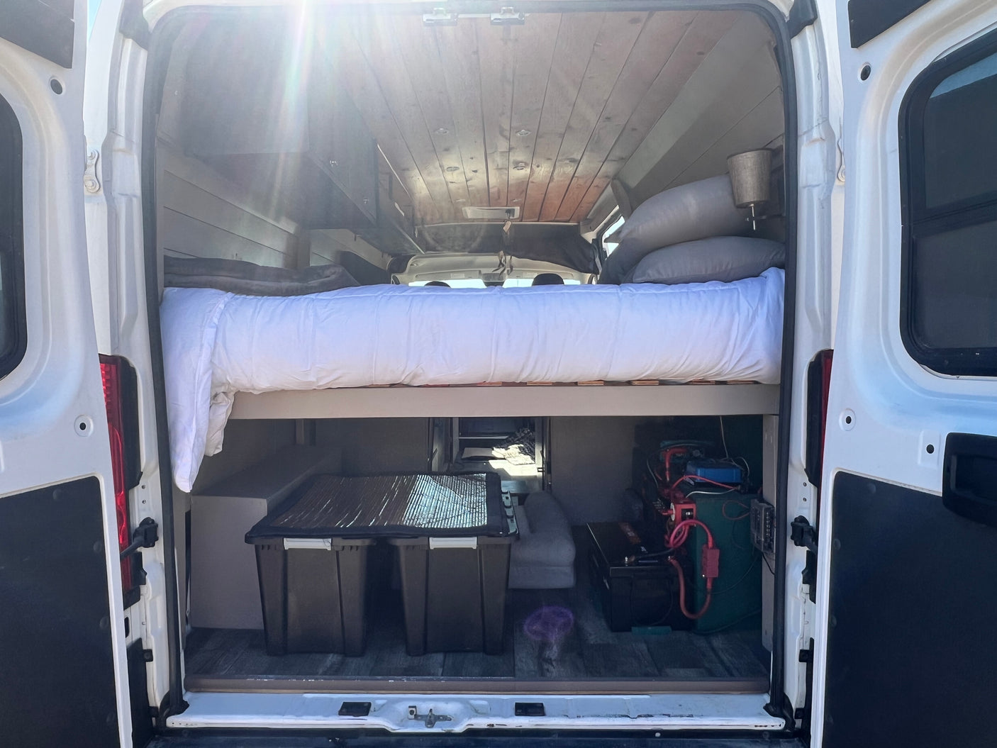 Brightly lit rv rental van interior: bed over storage bins & power setup. Wood ceiling & open rear doors.