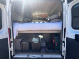 Brightly lit rv rental van interior: bed over storage bins & power setup. Wood ceiling & open rear doors.