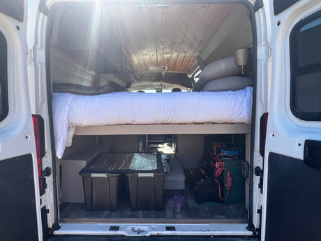 Brightly lit rv rental van interior: bed over storage bins & power setup. Wood ceiling & open rear doors.