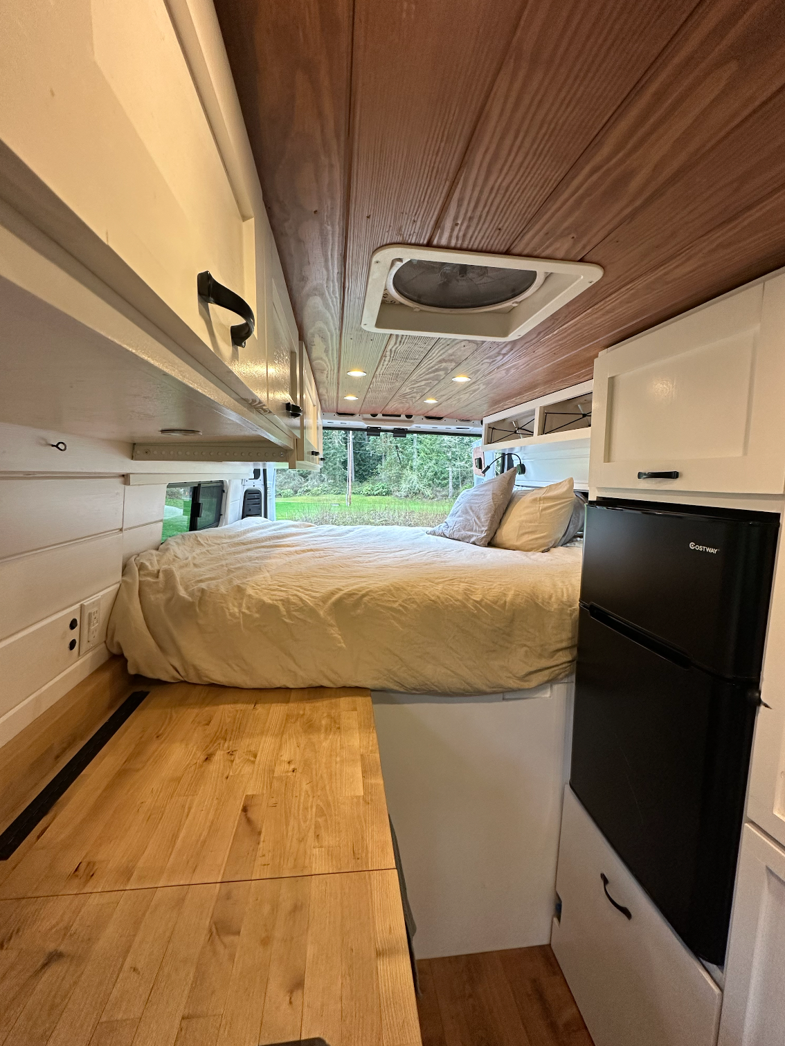 Cozy RV rental interior: bed, wood ceiling, white cabinets, compact fridge, and view to nature. Perfect for travel!