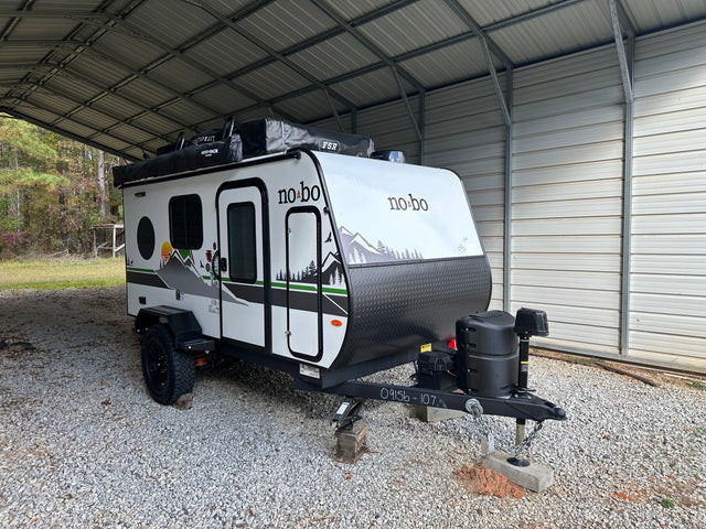 This no bo travel trailer, ready for your next outdoor rv rental adventure, is parked on gravel under a metal carport.