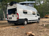 White Winnebago Solis RV rental camper van with pop-top at a rustic campsite. Logs, cords, and another unique RV are visible.