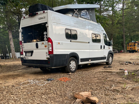 White Winnebago Solis RV rental camper van with pop-top at a rustic campsite. Logs, cords, and another unique RV are visible.