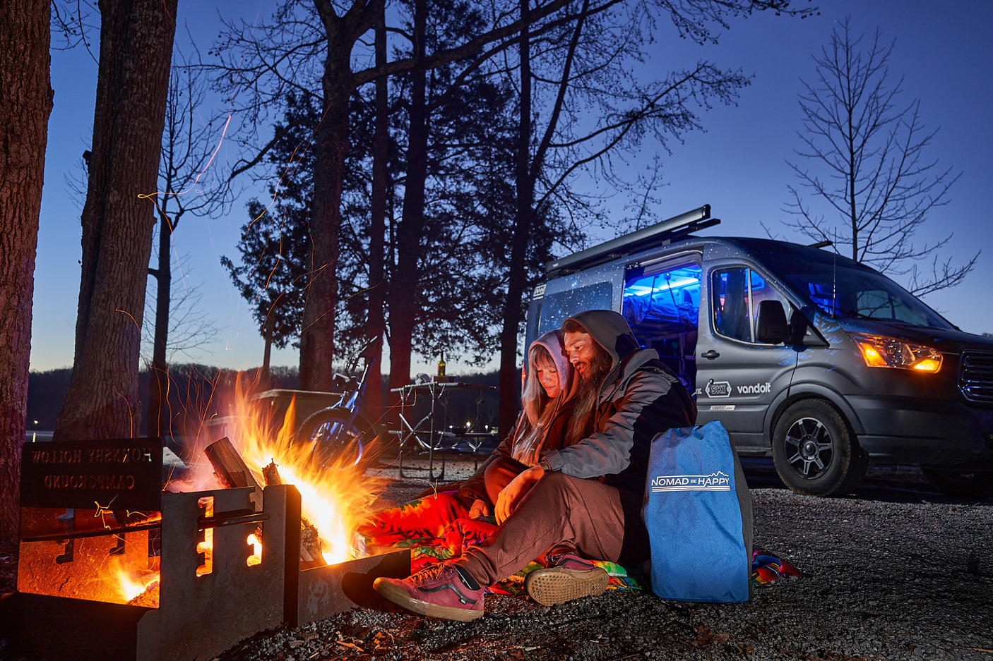 A couple enjoys a crackling campfire beside their RV rental camper van at a scenic lakeside campsite.