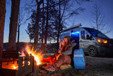A couple enjoys a crackling campfire beside their RV rental camper van at a scenic lakeside campsite.