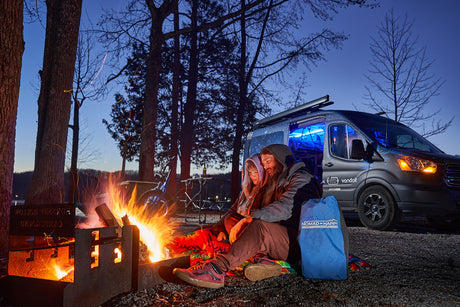 A couple enjoys a crackling campfire beside their RV rental camper van at a scenic lakeside campsite.