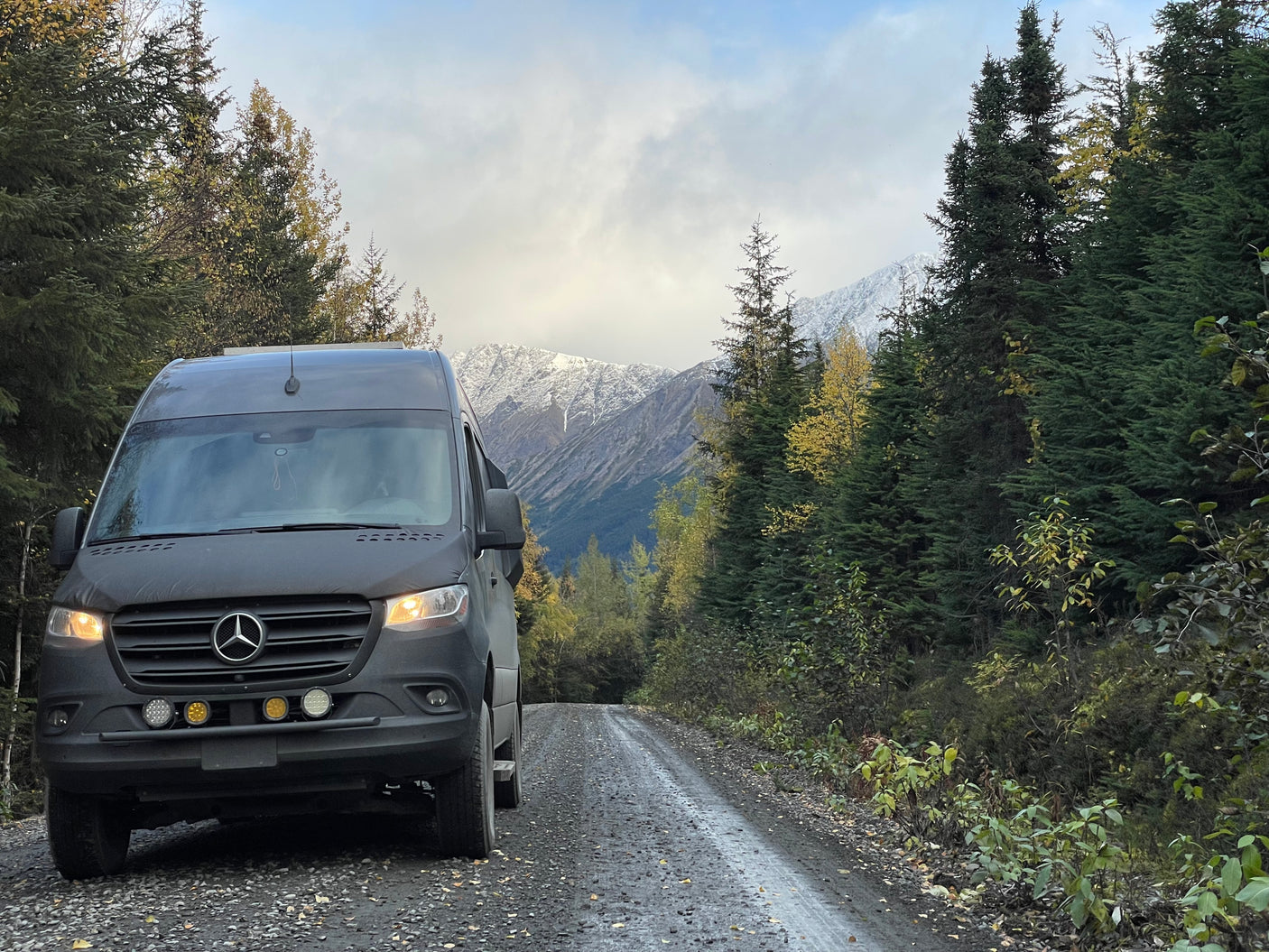 Mercedes-Benz Sprinter RV rental on a gravel road, surrounded by autumn trees and snowy mountains, perfect for adventure.