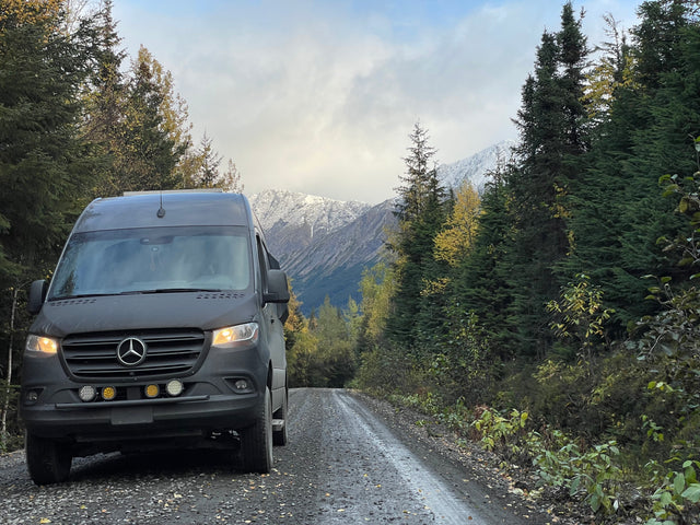 Mercedes-Benz Sprinter RV rental on a gravel road, surrounded by autumn trees and snowy mountains, perfect for adventure.