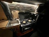 This RV rental's compact kitchen boasts a sink, two-burner stove with oven, and a fridge, all illuminated by LED strips for comfortable mobile living.