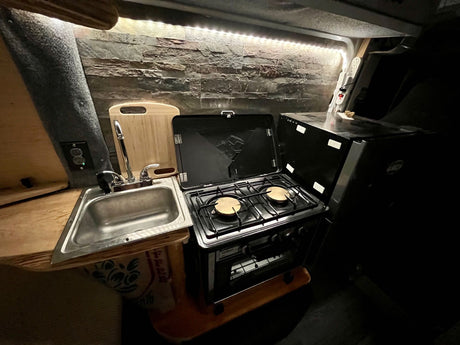 This RV rental's compact kitchen boasts a sink, two-burner stove with oven, and a fridge, all illuminated by LED strips for comfortable mobile living.