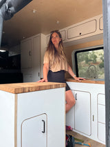 Woman enjoying a modern RV rental, showcasing its sleek interior, kitchen, and scenic window view. Ideal for travel!