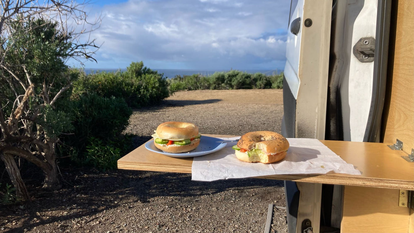 Ocean view RV rental breakfast: Two bagels on a wooden pull-out table outside a white van, surrounded by nature.