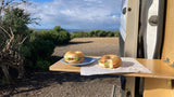 Ocean view RV rental breakfast: Two bagels on a wooden pull-out table outside a white van, surrounded by nature.