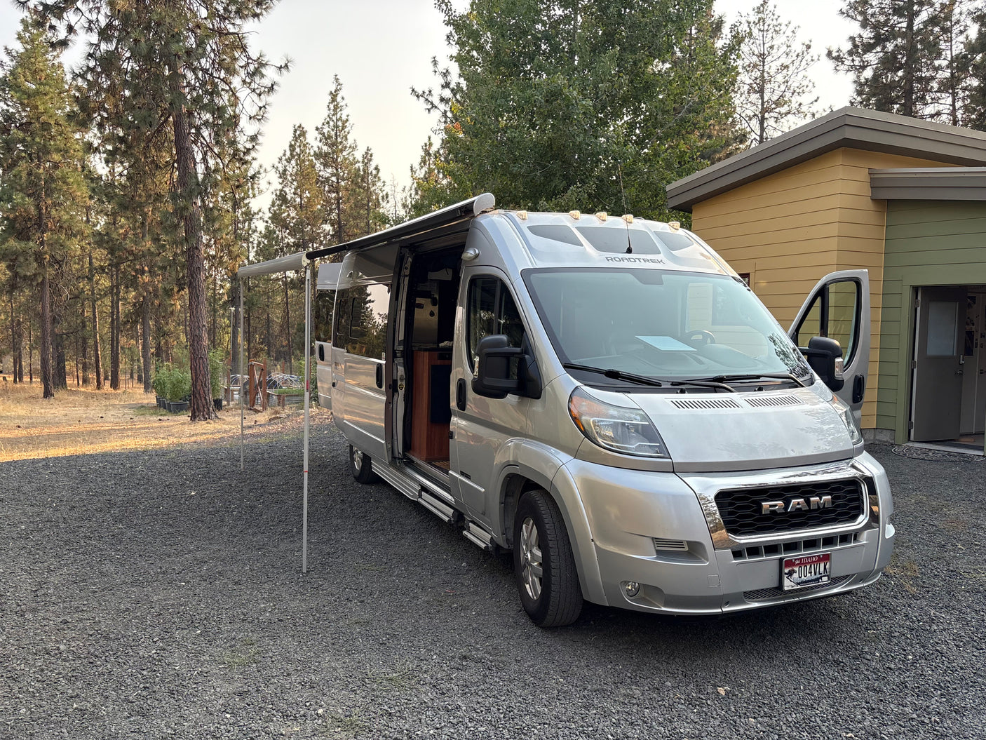 A silver Roadtrek RV rental, a type of camper van, parked on gravel with its awning extended in a natural, tree-filled setting.