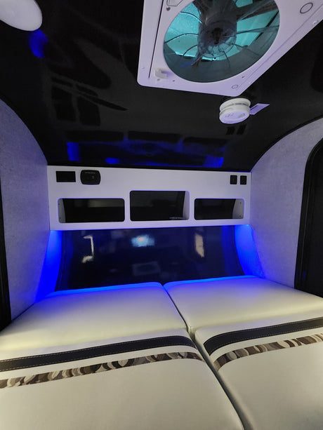 Modern RV rental interior featuring blue LED lighting, a comfortable bed, storage shelves, and a ceiling fan.