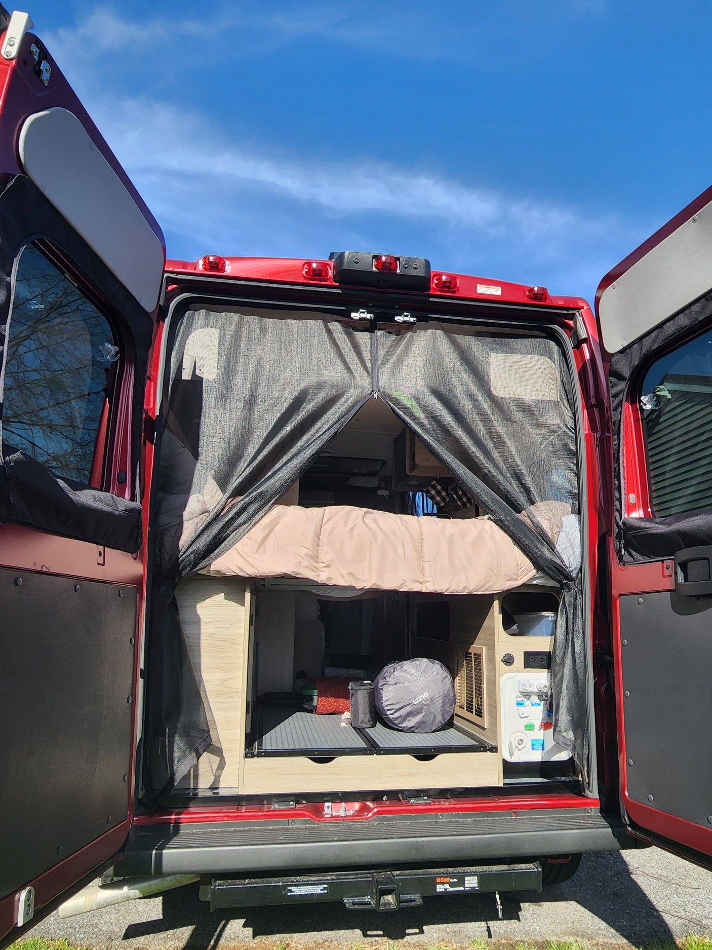 Open rear of a red camper van, ideal for an RV rental. Interior features a bed, control panel, and mesh screen.