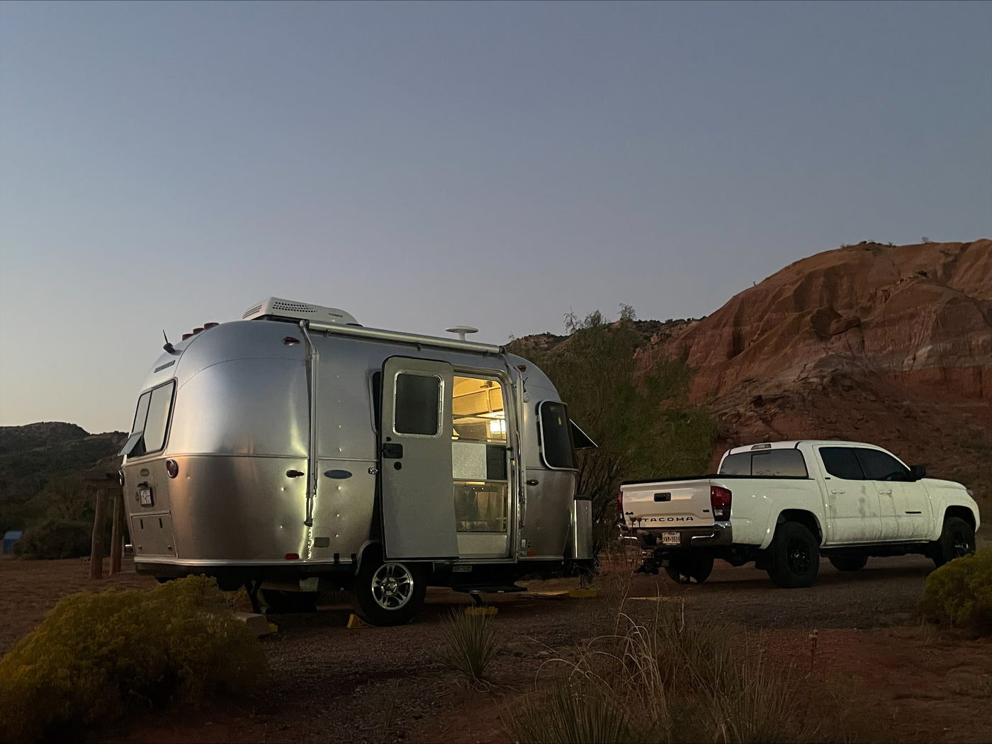 A shiny Airstream rv rental and white Tacoma truck parked in a desert landscape at dusk, with lights on inside the trailer. — photo 2