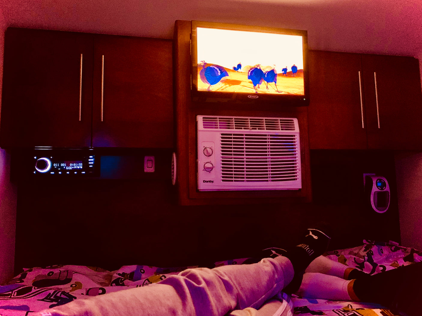 Cozy rv rental: feet up on bed, watching blue birds on TV. Amenities include AC, stereo, and overhead cabinets.
