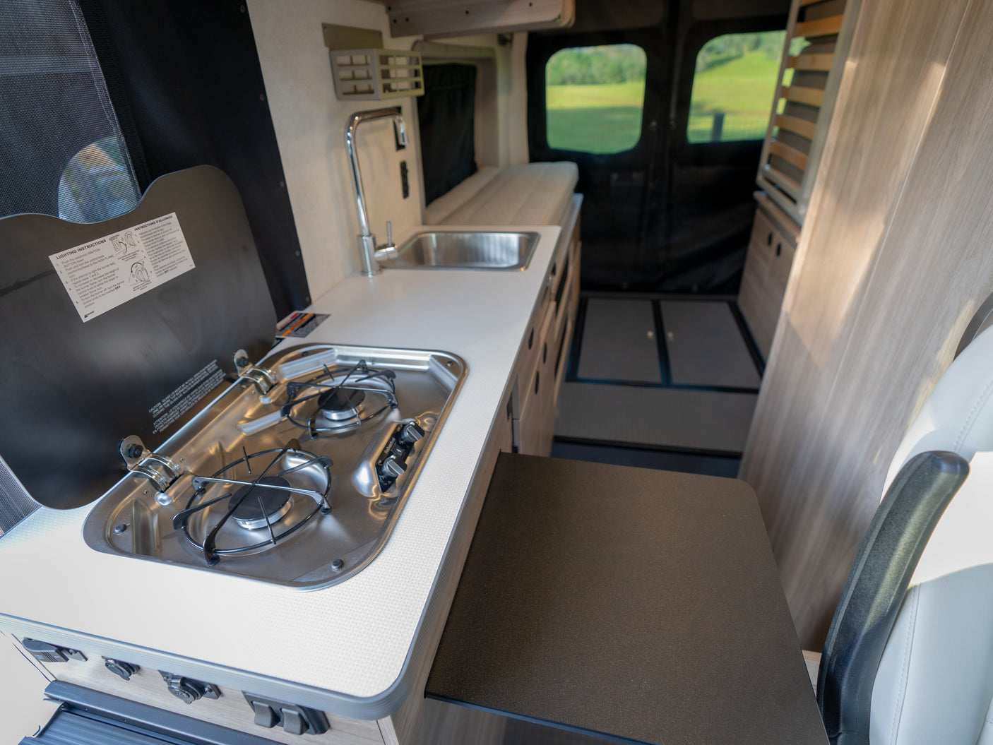 RV rental interior: A compact kitchen with a stove, sink, and counter offers a view of green fields outside the rear windows.