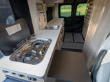 RV rental interior: A compact kitchen with a stove, sink, and counter offers a view of green fields outside the rear windows.
