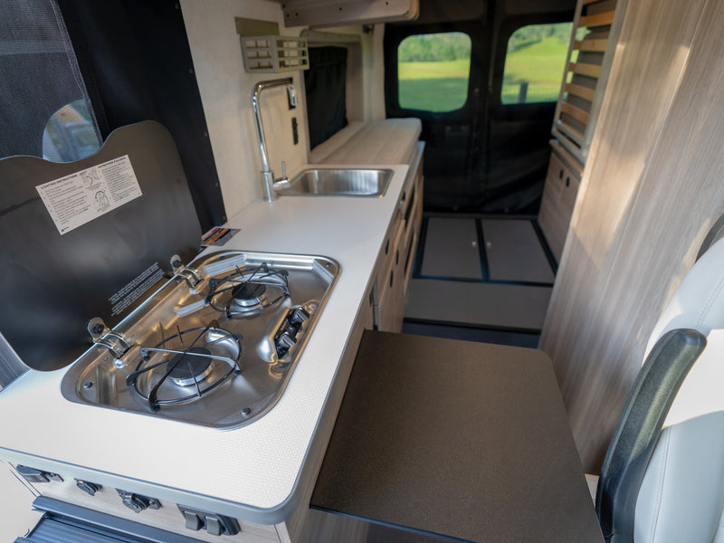 RV rental interior: A compact kitchen with a stove, sink, and counter offers a view of green fields outside the rear windows. — photo 14
