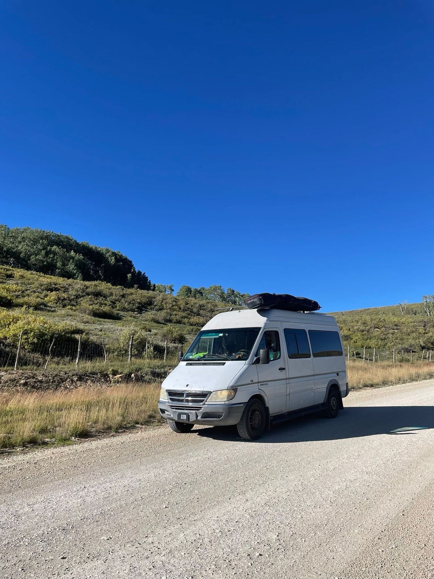  is parked on a scenic mountain road with open back doors and a person relaxing outside.|White camper van with roof cargo on a gravel road under a clear blue sky. Your ideal RV rental for outdoor adventures.|White RV rental — photo 3