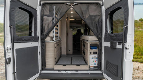 Open rear doors reveal an RV rental's camper van interior: bed, smart storage, and utility panel for comfortable mobile living.