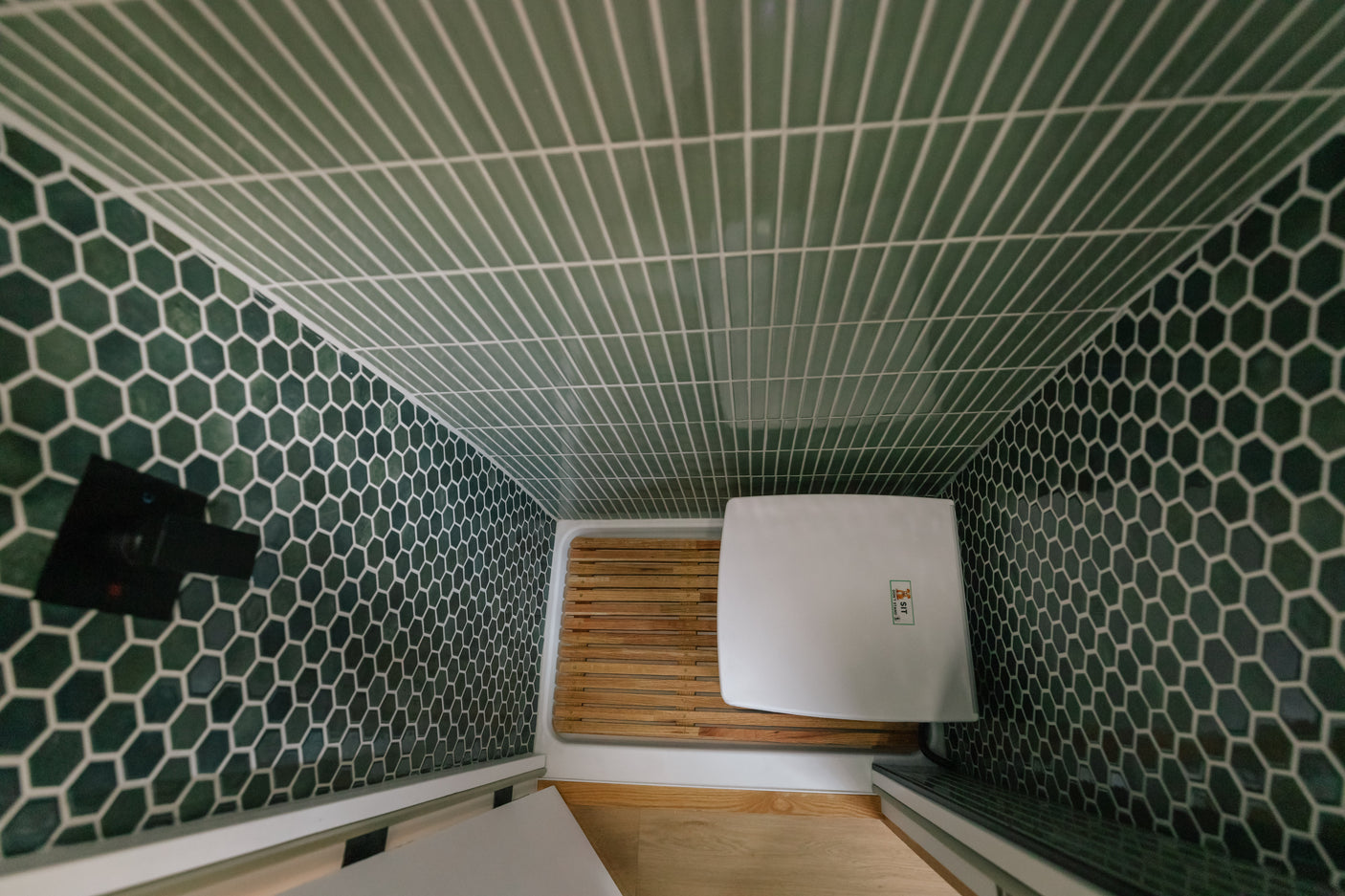 Inside an RV rental: a compact shower with green hexagonal & subway tiles, wooden mat, and a portable toilet.