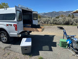 Scenic rv rental setup: a truck camper with open rear doors & pull-out storage, chairs, coolers, and mountains.