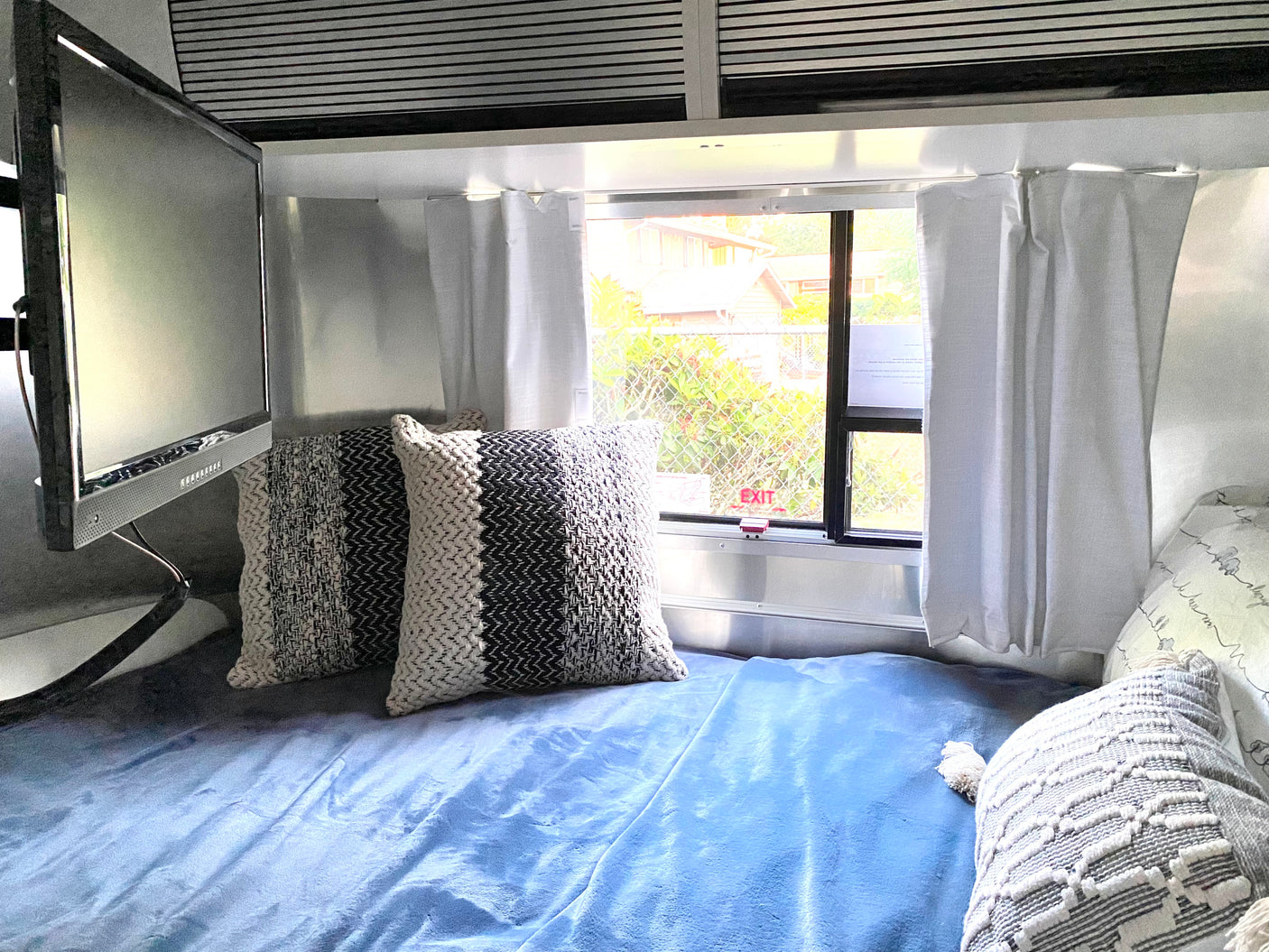A cozy RV rental bedroom features a blue blanket, patterned pillows, a wall-mounted TV, and a window with white curtains.
