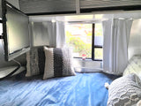 A cozy RV rental bedroom features a blue blanket, patterned pillows, a wall-mounted TV, and a window with white curtains.