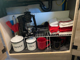 Organized RV rental cabinet with coffee/tea, mugs, lantern, utensils, and neatly folded towels, ideal for travel.